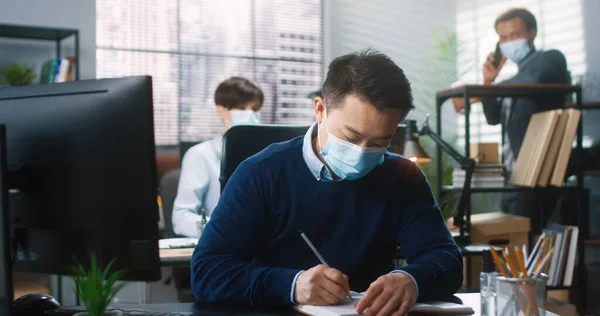 아시아 남성 노동자가 캐 비넷에 앉아 컴퓨터로 타이핑을 하고 플래너로 글을 쓰고 코로나 바이러스 격리 작업을 하는 모습. 뒤쪽에 동료들이 있어. 회사에서 의료용 마스크를 쓰고 일하는 사람 — 스톡 사진