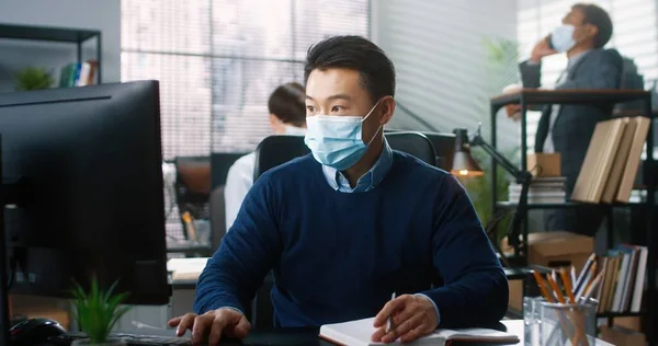 Retrato del trabajador asiático sentado en el escritorio en el gabinete escribiendo en la computadora y escribiendo en el planificador, el trabajo en cuarentena coronavirus. Colegas de fondo. Hombre en el trabajo de máscara médica en la oficina de la empresa — Foto de Stock