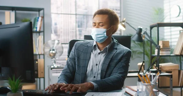 Retrato de un joven trabajador afroamericano con máscara médica sentado en el lugar de trabajo en el escritorio y escribiendo en la computadora mientras tose y tiene dolor de cabeza. pandemia de Coronavirus, síntomas de covid-19. Concepto de trabajo — Foto de Stock