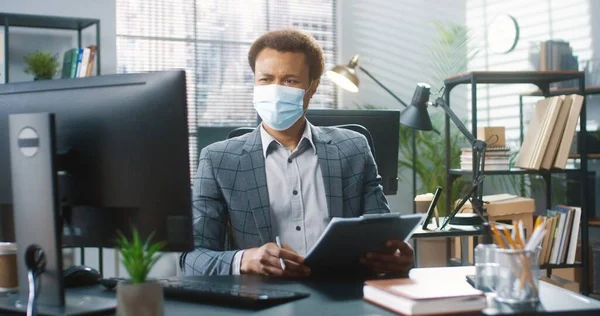 Retrato de hombre de negocios guapo afroamericano sentado en el escritorio en el gabinete escribiendo en la computadora y escribiendo en el planificador, el trabajo en cuarentena coronavirus. El hombre con máscara médica trabaja en la oficina de la compañía — Foto de Stock