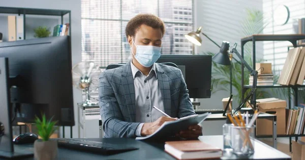 아프리카 계 미국인잘생긴 사업가가 캐 비넷에 앉아 컴퓨터로 타이핑을 하고 플래너로 글을 쓰고 코로나 바이러스 격리 작업을 하는 모습. 의료용 마스크를 쓰고 있는 사람 이 회사 사무실에서 일한다 — 스톡 사진