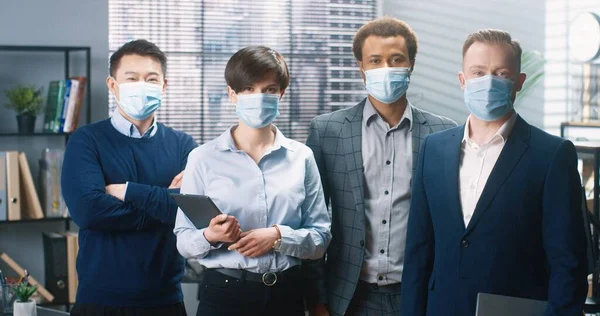 Retrato de jóvenes trabajadores mestizos alegres y positivos de pie en la oficina en compañía internacional, mirando a la cámara y sonriendo. Hombres y mujeres en máscaras médicas en el gabinete, concepto de negocio — Foto de Stock