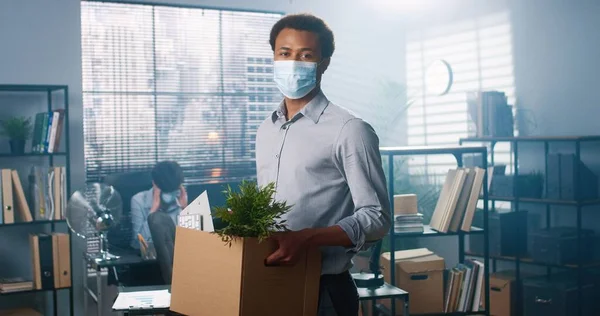 의료용 마스크를 쓴 젊은 아프리카계 미국 남성 이 상자를 손에 들고 캐비닛 안에서 서 물건을 들고 카메라를 보고 있는 모습, 직장에서 해고 된 사람, 사무실에서 일하는 신입 사원 — 스톡 사진