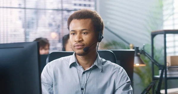 Close up retrato de feliz jovem afro-americano bonito operador de call center sentado na mesa no armário em fone de ouvido olhando para a câmera e sorrindo. Homem empregado no trabalho, conceito de trabalhador — Fotografia de Stock