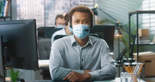행복하게 잘 생긴 아프리카 계 미국인 젊은이 가 사무실에서 책상에 앉아 컴퓨터로 타자를 치면서 의료용 마스크를 쓰고 있는 모습. 캐비닛에 있는 남성 콜 센터 운영자, 고객 지원 — 스톡 사진