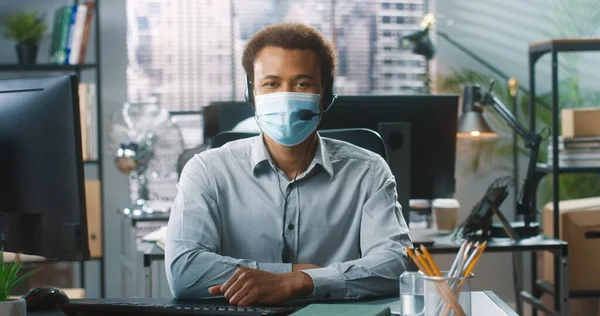 Retrato del joven afroamericano guapo feliz im máscara médica que trabaja mecanografiando en la computadora mientras está sentado en la mesa en la oficina. Operador de centro de llamadas masculino en auriculares en gabinete, soporte al cliente — Foto de Stock