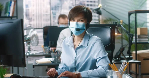 의료용 마스크를 쓴 젊은 백인 여성 이 사무실의 책상에서 연설하는 동안 컴퓨터로 타자를 치고 있는 모습. 중역 전화 캐비닛, 고객 지원 — 스톡 사진