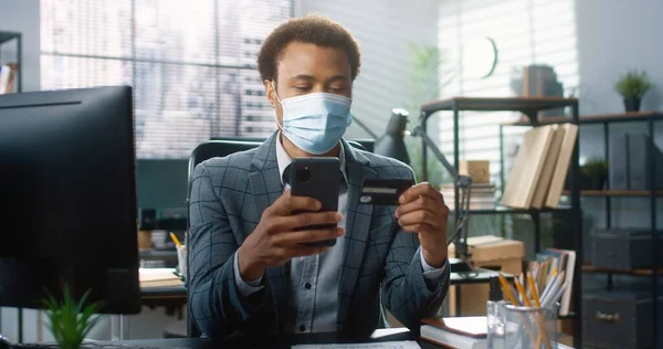 의료 마스크를 쓴 행복 한 아프리카 계 미국인 청년 이 책상에 앉아 스마트폰으로 온라인 도청을 하고 있습니다. 직장에서 인터넷 쇼핑을 하고 있습니다. 사업 컨셉, 초상화 — 스톡 사진