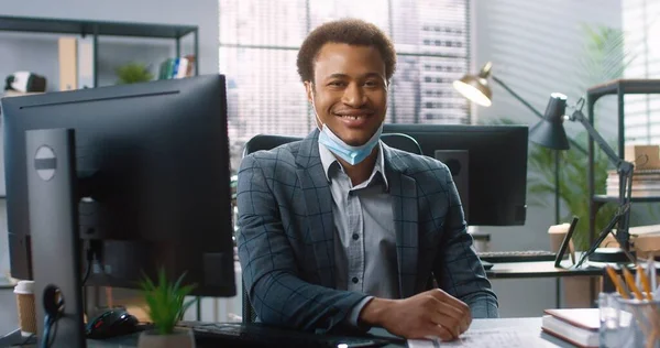 Porträtt av afroamerikansk stilig ung affärsman i medicinsk mask sitter vid bordet på arbetsplatsen, skriva på datorn surfa på nätet. Professionell i kabinettet. Arbetskoncept — Stockfoto