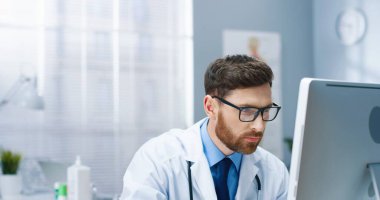 Gözlüklü beyaz önlüklü yakışıklı, konsantre olmuş beyaz sakallı erkek doktoru hastane dolabında oturmuş bilgisayar başında ekran monitörüne bakıyor ve okuyor. Çalışma konsepti