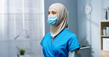 Tıbbi maskeli, üniformalı, hastanede kameraya bakan Arap genç ve güzel kadın pratisyen hekime yaklaş. Covid-19 salgını sırasında çalışan güzel kadın hemşire.