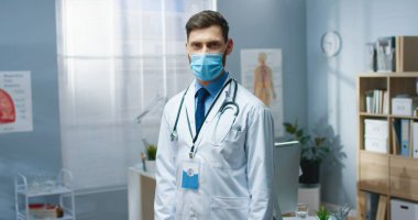 Tıbbi maskeli yakışıklı doktor portresi klinik dolabında tek başına kameraya bakıyor ve keyfi yerinde. Erkek genç doktor, karantina sırasında hastanede.