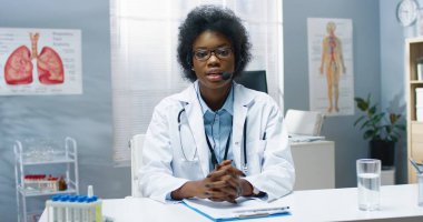 Neşe dolu, genç, Afro-Amerikalı kadın doktor portresi klinikte masada oturuyor ve kulaklıkla konuşuyor, tıbbi Webinar hastayla çevrimiçi video danışmanlığı öğretiyor.