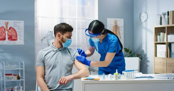 Junge hübsche asiatische Ärztin in medizinischer Maske und Schutzhandschuhen arbeitet im Krankenhaus und injiziert Coronavirus-Impfstoffe an einen hübschen kaukasischen männlichen Patienten. Covid Pandemie, Impfkonzept — Stockfoto