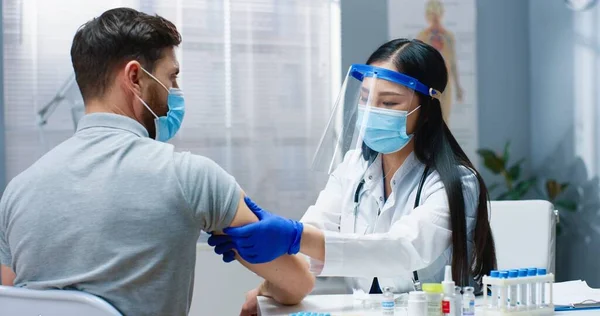 Крупный план портрета симпатичной азиатки-врача в медицинской маске и маске для лица, работающей в кабинете клиники во время вакцинации красивого белого пациента. Ковидская пандемия, концепция вакцинации — стоковое фото