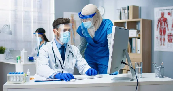 キャビネットで働く総合病院労働者の肖像。アラブの若い女性看護師と働く机の上に座ってコンピュータに入力する白人男性医師。医療、コロナウイルスのパンデミックの概念 — ストック写真