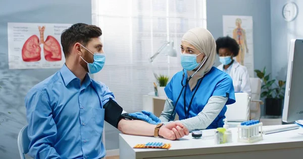 病院のキャビネットに座って白人男性患者への血圧を測定するかなり若いアラブ女性看護師。covid-19隔離中の医療センターの医療従事者。健康コンセプト — ストック写真