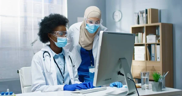 Портрет специалистов смешанной расы, работающих в кабинете в клинике. Арабская медсестра разговаривает с афроамериканским молодым врачом, печатающим в интернете. Концепция больницы — стоковое фото