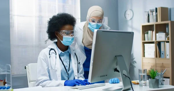 Retrato de especialistas en salud de raza mixta que trabajan en gabinete en clínica. Enfermera árabe hablando con médico joven afroamericano escribiendo en la navegación por computadora en línea. Concepto hospitalario —  Fotos de Stock