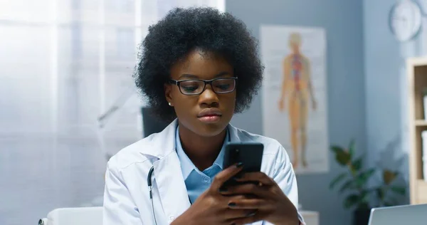 Nahaufnahme Porträt einer jungen, schönen afroamerikanischen Ärztin mit Brille, die im Krankenhaus sitzt und SMS auf ihrem Smartphone am Arbeitsplatz surft. Ärztin tippt auf Handy in Schrank — Stockfoto