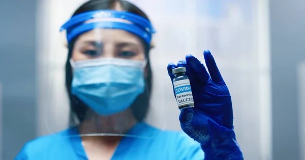Крупный план портрета азиатской молодой красивой медсестры в защитном щите и маске, держащейся за руки и показывающей вакцину Ковид-19, смотрящую в камеру. Врач проводит коронавирусное лечение, вакцинацию — стоковое фото