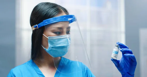 Крупный план азиатской молодой красивой женщины-врача в защитном щите лица и маске, держащейся за руку и показывающей ампулу вакцины Ковид-19, смотрящей в камеру. У медсестры коронавирусное лекарство — стоковое фото