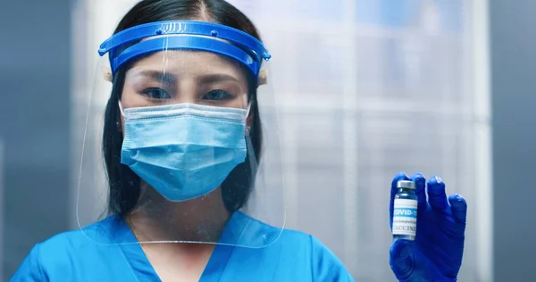 Крупный план молодой азиатской хорошенькой женщины-медика в защитном щите лица и маске, смотрящей на камеру, держащую в руке ампулу вакцины Ковид-19. Медсестра показала коронавирусную вакцинацию против пандемии — стоковое фото