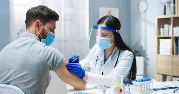 Крупным планом молодая азиатская красавица-врач в медицинской маске и со щитом на лице делает инъекцию вакцины ковид-19 в больницу красивому белому пациенту. Вакцинация, коронавирусное лечение — стоковое фото