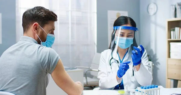 医療用マスクとフェイスシールド充填注射器で若いアジアのきれいな女性医師のクローズアップcovid-19ワクチンはハンサムな白人男性患者に病院で注射を行う。予防接種 — ストック写真