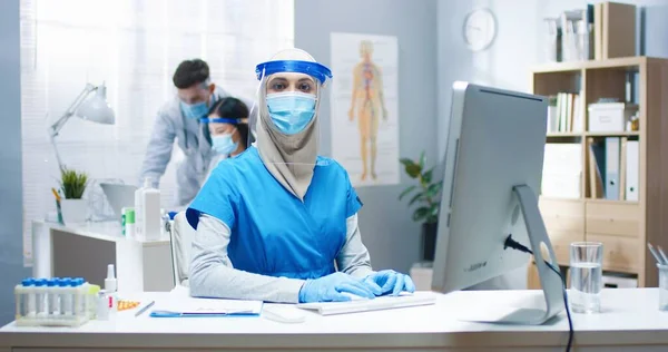 Портрет прекрасной молодой арабской женщины-врача в медицинской маске и маске для лица в больнице, сидящей за столом в кабинете и смотрящей в камеру. Женщина-врач работает с пандемией ковидов — стоковое фото