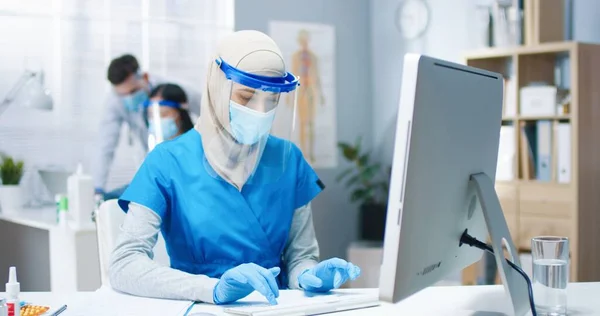 Retrato de bela jovem árabe ocupado enfermeira trabalhador de saúde em máscara médica e protetor facial sentado na mesa no gabinete digitando no computador que trabalha no hospital. Mulher médica, pandemia vívida — Fotografia de Stock