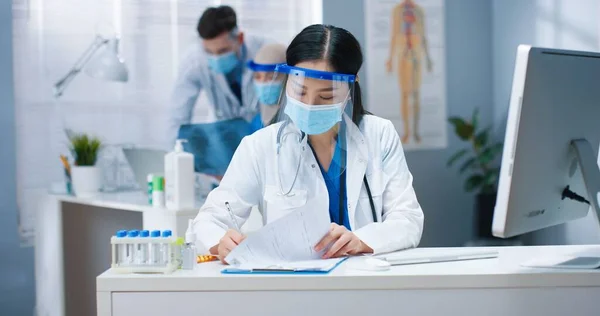 Портрет красивой молодой занятой азиатской женщины-медработницы в медицинской маске и щите на лице, сидящей за столом в кабинете, печатающей на компьютере и пишущей в документах. Больница, ковидная пандемия — стоковое фото