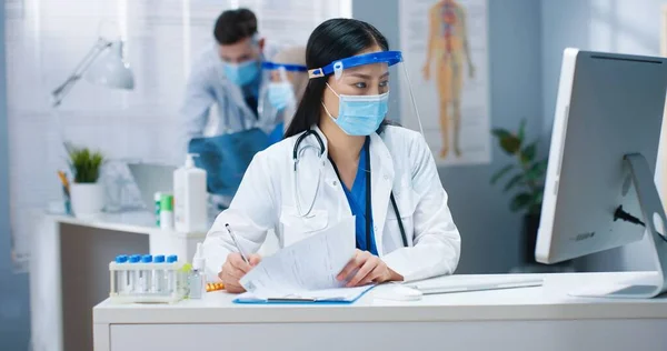 Porträt einer schönen jungen, geschäftigen asiatischen Gesundheitshelferin mit medizinischer Maske und Gesichtsschutz, die am Schreibtisch im Schrank sitzt, am Computer tippt und Dokumente schreibt. Krankenhaus, Covid Pandemie — Stockfoto