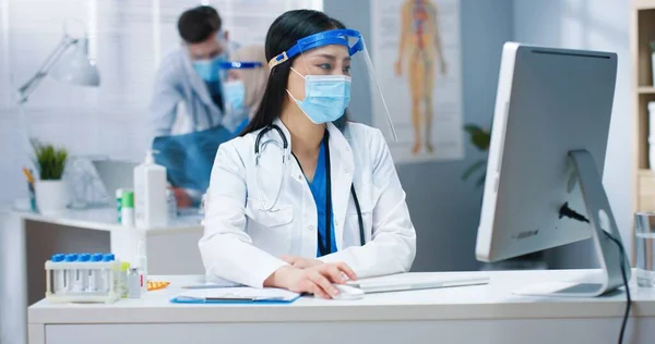 Портрет молодой серьезной азиатской женщины-терапевта в медицинской маске и маске для лица, сидящей за столом в белом халате и печатающей на компьютере, работающей в кабинете. Больница, коронавирусная пандемия, доктор — стоковое фото