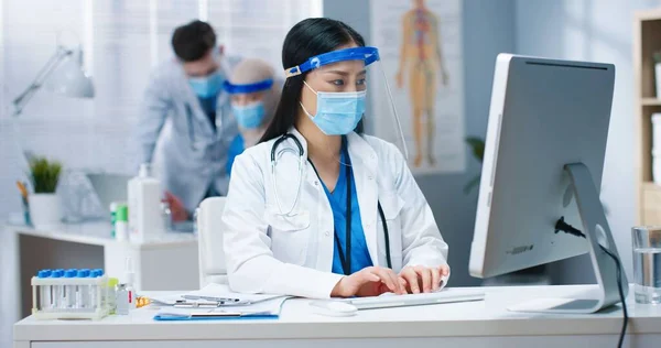 Retrato de jovem séria asiática praticante geral em máscara médica e protetor facial sentado na mesa em casaco branco digitando no computador trabalhando no gabinete. Hospital, pandemia de coronavírus, médico — Fotografia de Stock