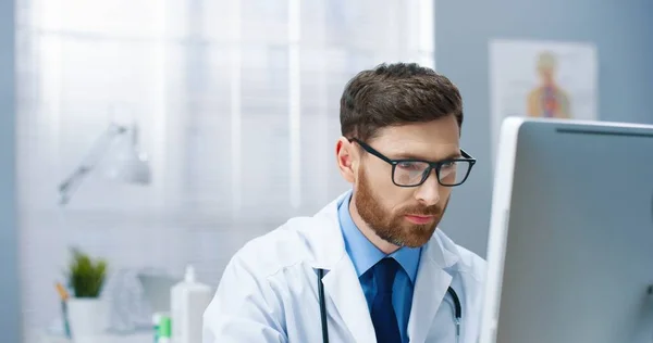 Großaufnahme eines gutaussehenden jungen kaukasischen bärtigen Arztes im weißen Arztkittel mit Brille, der im Krankenhausschrank sitzt, am Computer arbeitet, auf den Bildschirm schaut und liest. Gesundheitswesen, Klinikkonzept — Stockfoto