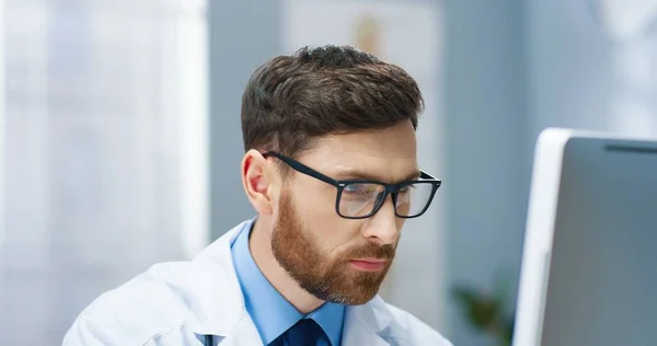 Großaufnahme eines gutaussehenden jungen kaukasischen bärtigen Arztes in weißem Mantel mit Brille, der im Krankenhausschrank sitzt, am Computer arbeitet, auf den Bildschirm blickt und liest. Arbeitskonzept — Stockfoto