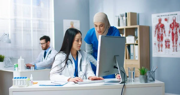 Schöne gemischte Gesundheitsspezialisten, die am Arbeitsplatz sprechen. Asiatische junge Ärztin surft am Computer und spricht mit arabischen Mitarbeiterinnen. Klinik, medizinisches Konzept — Stockfoto