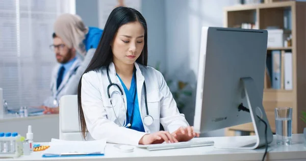 Porträt einer schönen jungen asiatischen Ärztin in weißem Arztkittel, die am Schreibtisch am Arbeitsplatz im Krankenhausschrank sitzt und die Ergebnisse der Leseanalyse schreibt. Krankenpfleger in Klinik — Stockfoto