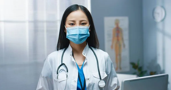 Крупный план азиатской молодой красивой женщины-терапевта в медицинской маске и белом халате, стоящей в больнице, смотрящей в камеру и улыбающейся в хорошем настроении, работающей во время пандемии коронавируса — стоковое фото