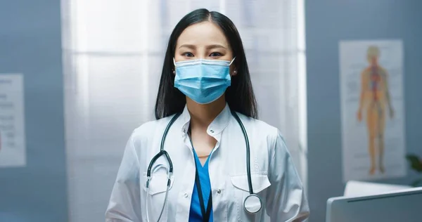 亚洲年轻貌美的全科医生，戴着医疗面罩，身穿白衣，站在医院里，看着相机，带着愉快的笑容，在大脑皮层病毒大流行期间工作 — 图库照片