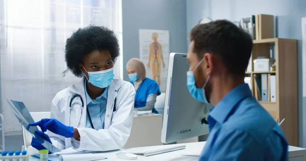 Nahaufnahme einer afroamerikanischen Ärztin in medizinischer Maske, die im Krankenhausschrank sitzt und mit einem jungen, gut aussehenden Mann aus dem Kaukasus spricht, der die Röntgenuntersuchung erklärt. Coronavirus — Stockfoto
