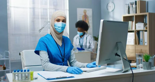 幸せなアラビア語の肖像かなり若い女性のプロのセラピスト職場でのコロナウイルス検疫キャビネットで良い気分でカメラを見てテーブルに座っている。病院、診療所のコンセプト — ストック写真