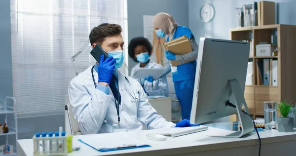 Retrato de branco bonito jovem médico do sexo masculino falando no smartphone com o paciente olhando para a tela do computador discutindo resultados de análise. Trabalho em covid-19 pandemia, conceito de hospital — Fotografia de Stock