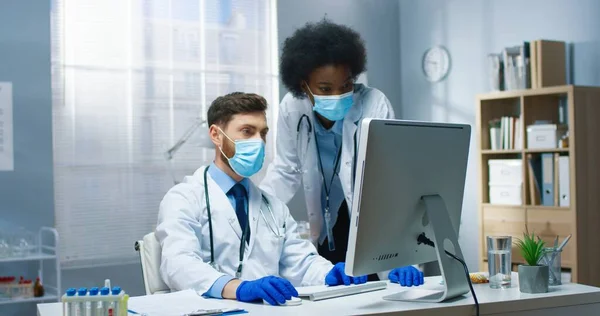 Mitarbeiter multiethnischer Ärzte in medizinischen Masken sprechen im Kabinett in der Klinik und blicken auf den Computerbildschirm. Schöner kaukasischer Mann tippt auf Computer und spricht mit afroamerikanischer Ärztin — Stockfoto