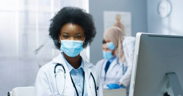 Primer plano retrato de alegre joven bonita mujer afroamericana en máscara médica que trabaja en la computadora mirando pantalla del monitor sentado en el gabinete en el hospital. pandemia de Covid-19, atención sanitaria —  Fotos de Stock