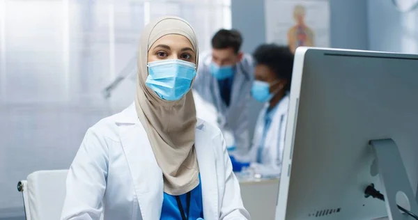 Nahaufnahme Porträt der schönen arabischen jungen Ärztin, die am Arbeitsplatz am Schreibtisch im Kabinett sitzt und in die Kamera blickt. Arbeiten während der covid-19-Pandemie. Krankenhaus, Klinikkonzept — Stockfoto