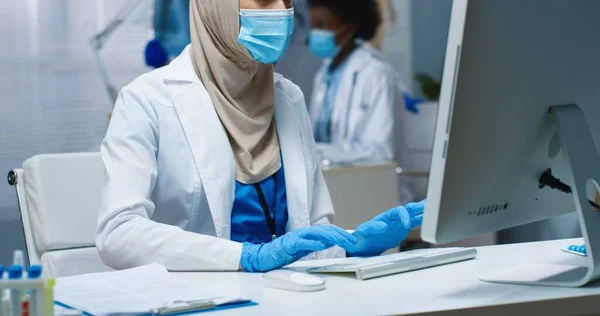 病院のタイピングやコンピュータ上でオンライン閲覧でキャビネットで働く医療用マスクのかなり若いアラビア人女性医師の閉鎖。Covid-19パンデミック、医療、医療センターの概念 — ストック写真