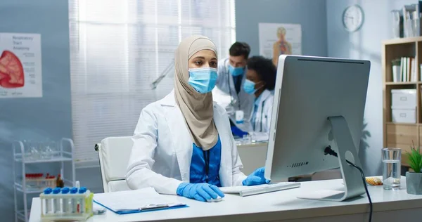 Молоді арабські прекрасні жінки-лікар в медичній масці працюють, друкують і переглядають онлайн на комп'ютері сидячи на робочому місці. Вірус Ковід-19, охорона здоров'я, медична концепція — стокове фото