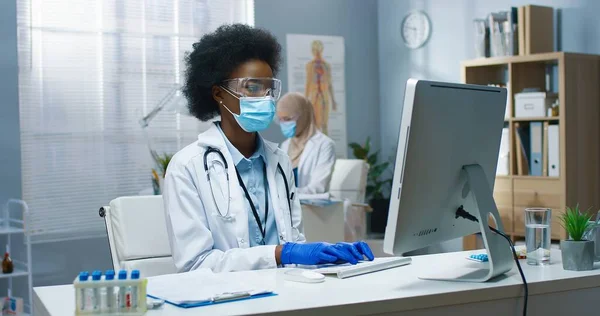 Junge afroamerikanische schöne professionelle Ärztin mit medizinischer Maske und Brille arbeitet, tippt und surft online am Computer am Arbeitsplatz. Covid-19 Virus, medizinisches Konzept — Stockfoto